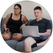 A young man and a young woman in glasses sit on a dark gray couch. The man is using a laptop.
